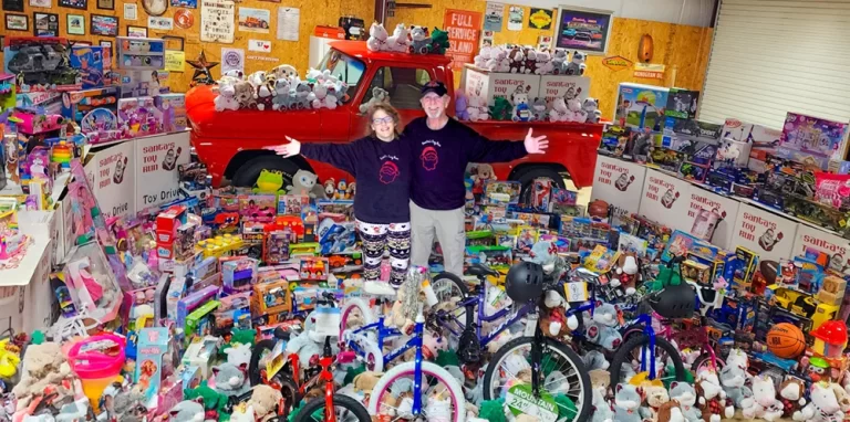 Two volunteers with arms wide in front of a red pickup ringed by toys and bikes